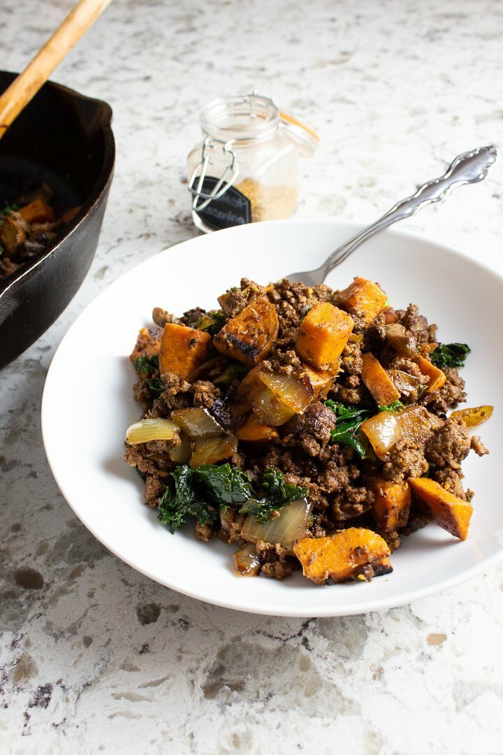 Breakfast hash with ground beef, onions, yam, mushrooms and kale served up on a white plate.